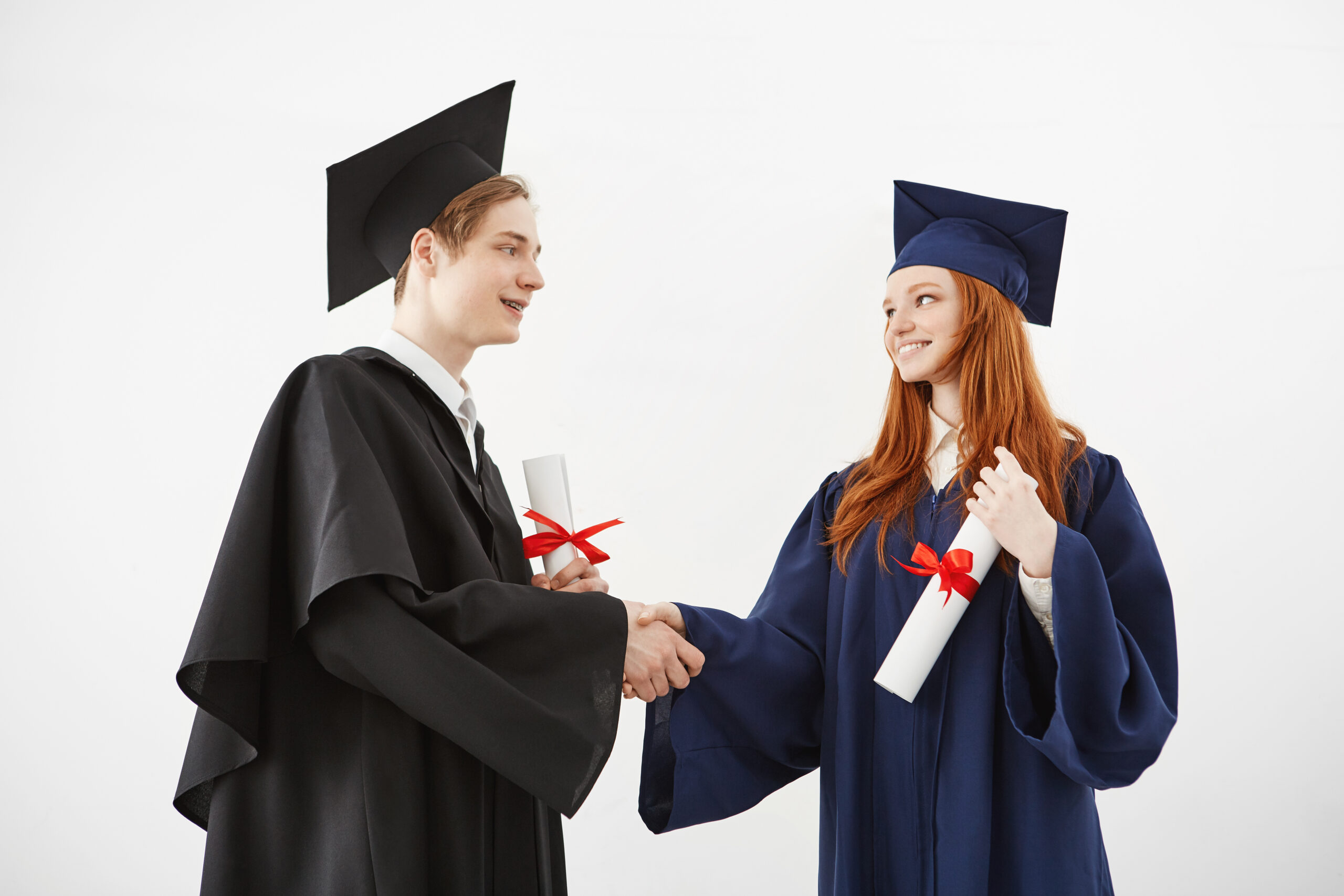Two graduates classmates shake hands smiling holding diplomas over white background. Two graduates classmates in mantles shake hands smiling holding diplomas over white background.